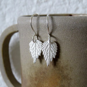 May include: A pair of silver earrings featuring a delicate leaf design. The leaves are textured and have a slightly raised appearance. The earrings are hanging from a simple hook.