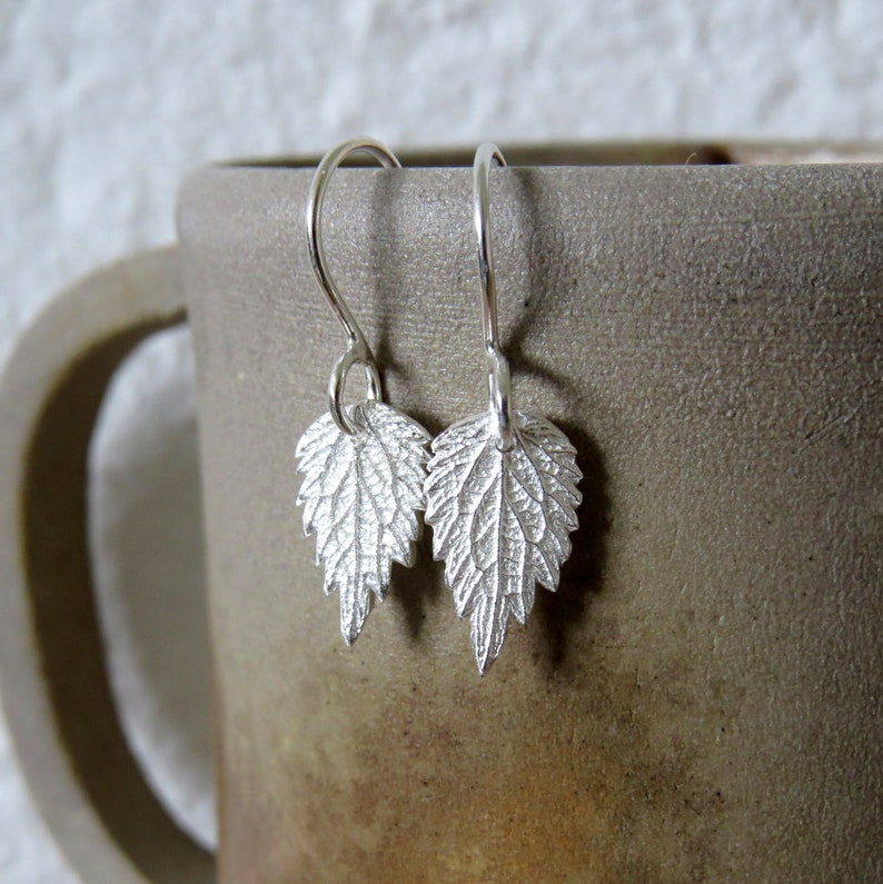 May include: A pair of silver earrings featuring a delicate leaf design. The leaves are textured and have a slightly raised appearance. The earrings are hanging from a simple hook.