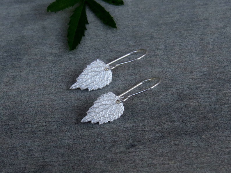 May include: A pair of silver earrings shaped like leaves. The leaves have a textured surface and are attached to silver hooks.