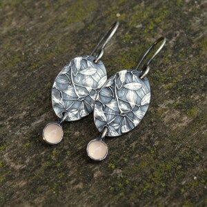 May include: A pair of silver-toned oval earrings with a leaf design. Each earring has a small, round, pale pink stone dangling from the bottom. The earrings are displayed on a textured, green-toned surface.