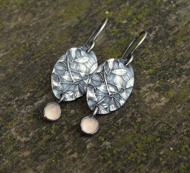 May include: A pair of silver-toned oval earrings with a leaf design. Each earring has a small, round, pale pink stone dangling from the bottom. The earrings are displayed on a textured, green-toned surface.