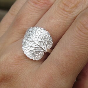 May include: A silver ring with a detailed leaf design. The ring is worn on a person's finger.