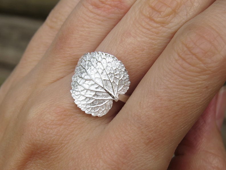 May include: A silver ring with a detailed leaf design. The ring is worn on a person's finger.