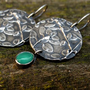 Puede incluir: Un par de pendientes de plata con un dise&ntilde;o floral y detalles de piedras preciosas verdes. Los pendientes est&aacute;n hechos de metal texturizado y tienen un aspecto r&uacute;stico y hecho a mano.