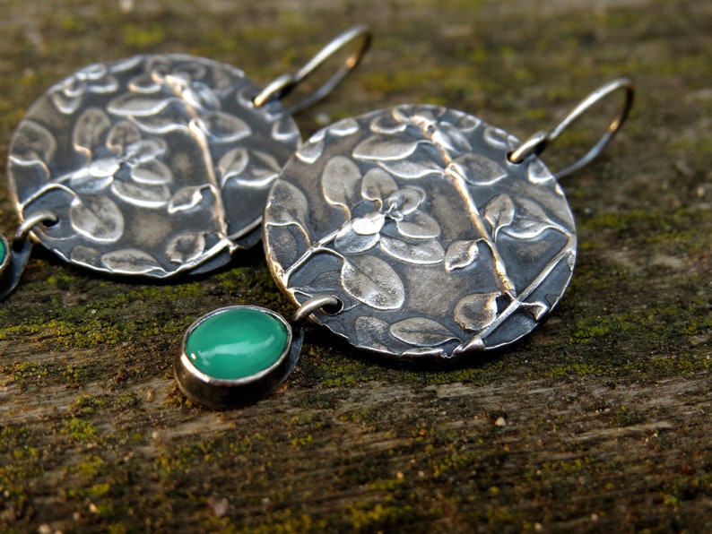 Puede incluir: Un par de pendientes de plata con un dise&ntilde;o floral y detalles de piedras preciosas verdes. Los pendientes est&aacute;n hechos de metal texturizado y tienen un aspecto r&uacute;stico y hecho a mano.