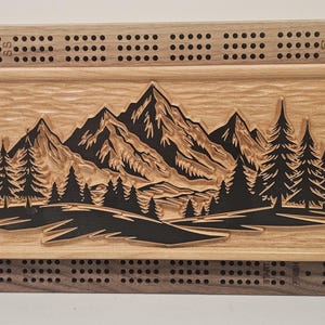 Può includere: Tavolo da cribbage in legno con un paesaggio di montagne e foreste intagliato al centro. La tavola è incorniciata con un legno più scuro e presenta fori per i pioli. La scena della montagna è dettagliata con alberi e vette.