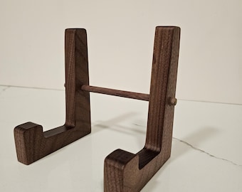 May include: A dark brown wooden display stand with a horizontal bar. The stand has a rectangular base with a curved top and a vertical support. The stand is designed to hold a plate or other item.