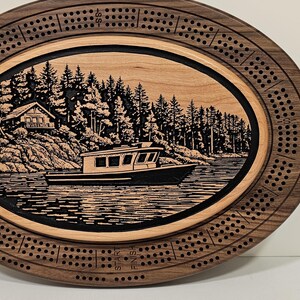 Può includere: Una tavola da cribbage ovale in legno con un anello esterno in noce scuro con fori per il punteggio e un intarsio centrale in legno chiaro. L'intarsio raffigura una scena incisa in nero di una barca sull'acqua vicino a una baita e pini.
