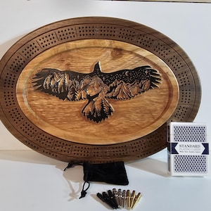 May include: An oval wooden cribbage board featuring a carved eagle design. The eagle has outstretched wings, mountains, and a starry night sky. Includes a deck of playing cards, pegs, and a black velvet bag.
