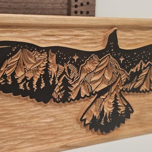 May include: A wooden carving of an eagle in flight, with a detailed mountain and starry night scene within its wings. The eagle is carved from light-colored wood, with the mountain scene in black. The carving is rectangular.