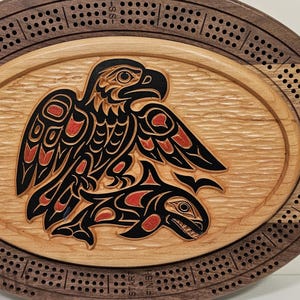 Op de afbeelding: Een ovaal houten cribbagebord met een donkere walnoothouten buitenring met boorgaten voor de puntentelling en een lichter esdoornhouten middenpaneel. Het midden toont een uitgesneden adelaar- en orka-ontwerp in zwart en rood, met START en FINISH.
