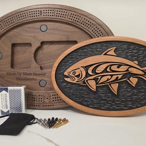 May include: A wooden cribbage board set with a carved fish design. The set includes a dark wood oval cribbage board, a deck of playing cards, pegs, and a black drawstring bag. The cribbage board has the text "Made by Black Beaver Woodworks".