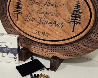May include: A wooden cribbage board with an oval center, engraved with the words "Gary & Terri, New Home, New Beginning, New Memories, EST 2022." Includes pegs, a black velvet bag, and a deck of cards.