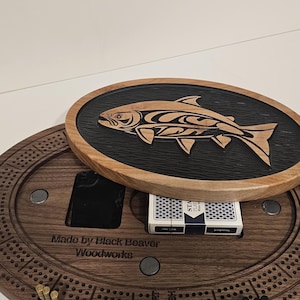 Puede incluir: Tablero de cribbage de madera con un diseño decorativo de pez en la tapa. El tablero es de color marrón oscuro con una pista circular y agujeros para clavijas. Incluye una baraja de cartas y clavijas. La tapa tiene un tallado de pez.