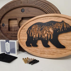 Peut inclure: Ensemble de jeu de société en bois avec un motif d'ours, un jeu de cartes et des chevilles. Le motif de l'ours est sculpté dans le bois et rempli d'une teinture noire. L'ensemble comprend un sac en velours noir et un jeu de société ovale marron foncé.