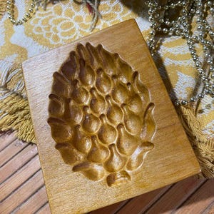 May include: A rectangular wooden mold with a detailed pinecone design carved into its surface. The mold is a warm, golden brown color, and the pinecone is intricately detailed. The mold is likely used for baking or crafting.