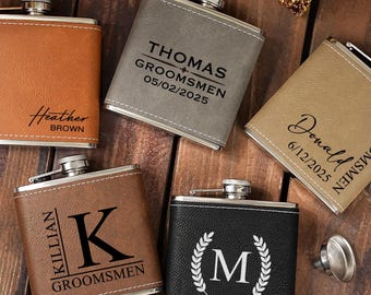 May include: An assortment of personalized flasks in various colors, including brown, gray, and black. Each flask features custom text, such as names, dates, and initials, making them ideal for groomsmen gifts. The flasks are displayed on a wooden surface.