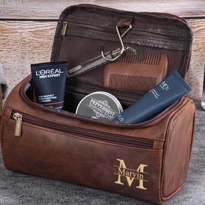 May include: A brown leather toiletry bag with a zipper, containing grooming products. The bag is personalized with the name "Marvin" and the initial "M". Other items include a razor, comb, and skincare products.