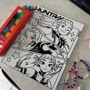 Pode incluir: Uma impressão em tela em preto e branco em estilo de banda desenhada com três figuras femininas estilizadas, o texto "HUNTR" e nomes de personagens. Uma caixa vermelha de tintas aquarela e pulseiras de contas também são visíveis.