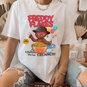 Puede incluir: Camiseta blanca con un gráfico de Freddy Krueger sosteniendo una cuchara sobre un tazón de cereal. El texto "FREDDY FLAKES" está encima de la imagen, con "A Nightmare You Can CRUNNCH!" debajo. Un logo de "FACE RAZOR" está a la izquierda.