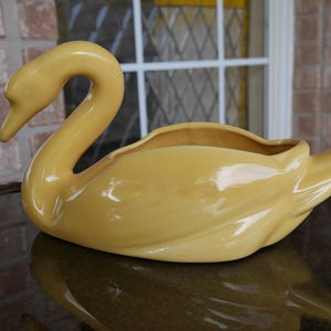 May include: A yellow ceramic swan planter with a smooth, glossy finish. The swan's neck is curved and the planter has a wide, open mouth.