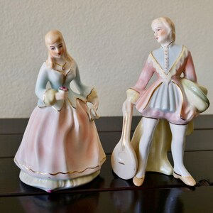 May include: Two porcelain figurines of a man and woman in period clothing. The woman wears a light blue and pink dress with gold trim, holding a small flower. The man has a pink and blue outfit and holds a lute. The figurines are on a dark surface.