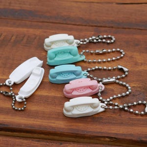 May include: A collection of colorful, miniature telephone keychains in white, teal, light blue, and pink. Each keychain features a small, retro-style phone charm attached to a silver ball chain. The background is a dark wooden surface.