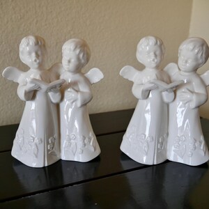 May include: Two sets of white ceramic angel figurines. Each set features two angels holding a book and wearing robes with floral detailing. The angels have small wings and are standing on a black surface.