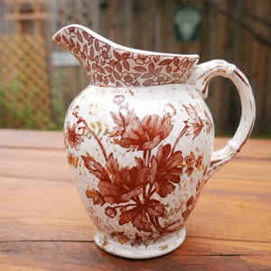 May include: A vintage ceramic pitcher with a white base and a brown floral pattern. The pitcher has a spout, a handle, and a decorative floral design around the rim. The pitcher is likely used for serving beverages.