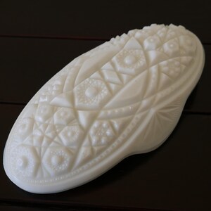 May include: White milk glass dish with a raised geometric pattern. The dish is oval shaped and has a smooth, glossy finish.