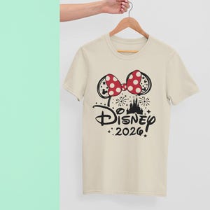 May include: A cream-colored t-shirt featuring a Minnie Mouse-inspired design. The design includes a red polka-dot bow, black mouse ears, and the word "Disney" above "2026!" in black, with a castle silhouette and fireworks.