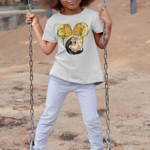May include: A young girl wearing a white t-shirt with a graphic of a yellow bow and a cartoon character. She is wearing gray sweatpants and pink sneakers. She is on a swing set.