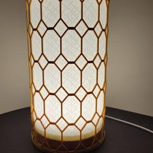 Puede incluir: Lámpara de mesa cilíndrica con un patrón geométrico de panal. La lámpara emite una luz cálida y brillante a través de la pantalla blanca estampada. El marco es de color marrón cálido.