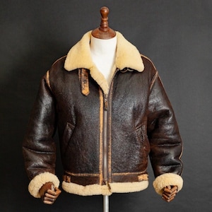 May include: A brown leather bomber jacket with a cream shearling collar and cuffs. The jacket features a zip-up front, two front pockets, and a brown leather strap detail on the collar. The jacket is displayed on a wooden mannequin.