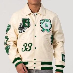Könnte beinhalten: Cremefarbene Collegejacke mit grünen und schwarzen Aufnähern. Verfügt über einen großen grünen "B" Aufnäher, einen Geldsack, Diamantenformen und die Zahl "33". Die Jacke hat grün-weiß gestreifte Bündchen und Saum.
