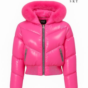 Puede incluir: Chaqueta acolchada rosa brillante con capucha ribeteada de piel. La chaqueta tiene un acabado brillante, una cremallera negra y dos bolsillos con cremallera. Los puños y la cintura son de canalé. El texto "GOLD STAR SKT" es visible en la esquina superior derecha.