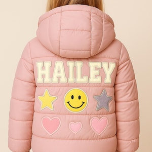 May include: A pink puffer jacket with a hood, displaying the name "HAILEY" in cream-coloured letters on the back. The jacket is adorned with a yellow star, a smiley face, a grey star, and pink hearts. The text "Gold Star Skt" is visible.