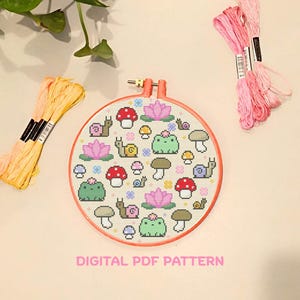 Op de afbeelding: Een ronde borduurring met een kruissteekpatroon van groene kikkers, roze lotusbloemen, rood-witte paddenstoelen en slakken. De tekst DIGITAL PDF PATTERN staat onder de ring, geflankeerd door geel en roze borduurgaren.