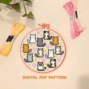 Op de afbeelding: Een borduurring met een kruissteekpatroon van gepixelde katten in verschillende kleuren. Roze en geel borduurgaren zijn rond de ring geplaatst. De tekst "DIGITAL PDF PATTERN" wordt eronder weergegeven.