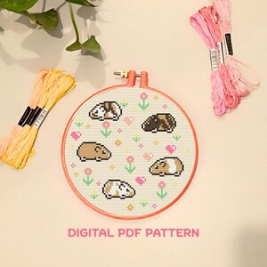 May include: A cross-stitch pattern in a round hoop featuring pixelated guinea pigs in various colors, surrounded by flowers and hearts. The hoop is coral-colored, with bundles of pink and yellow embroidery floss. Text at the bottom reads "DIGITAL PDF PATTERN."
