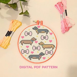 Op de afbeelding: Een kruissteekpatroon in een borduurring met teckels, roze strikken en hartjes. De tekst "DIGITAL PDF PATTERN" staat eronder. Roze en gele borduurgaren zijn zichtbaar.