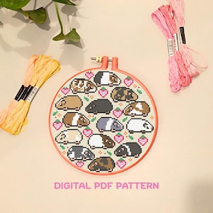 Op de afbeelding: Een kruissteekpatroon met meerdere cavia's in verschillende kleuren, omringd door roze hartjes, in een ronde oranje borduurring. De tekst "DIGITAL PDF PATTERN" staat eronder. Bundels roze en gele borduurgaren zijn ook aanwezig.