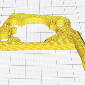 Könnte beinhalten: Ein gelbes 3D-gedrucktes Kunststoffteil mit einem großen Loch in der Mitte und einem kleineren Loch an der Seite. Das Teil hat eine lange, dünne Verlängerung auf der rechten Seite.