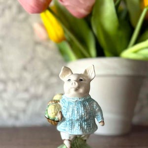 May include: A ceramic pig figurine wearing a light blue sweater and carrying a basket of yellow objects. The pig stands on a patch of green grass. Pink and yellow tulips are in the background.