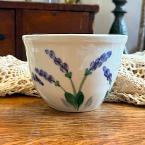May include: A white ceramic bowl featuring a hand-painted design of purple lavender sprigs with green stems and leaves. The bowl has a smooth, rounded shape with a slightly flared rim, showcasing a botanical motif on its exterior surface.