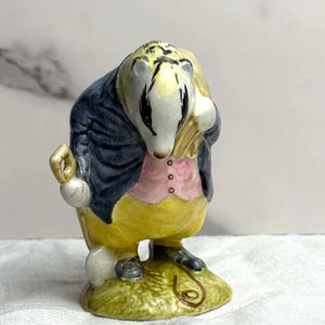 May include: A ceramic figurine of a badger dressed in a blue coat, yellow vest, and pink shirt. The badger is holding a bag and a gold-colored object. It stands on a green base with a brown swirl design.