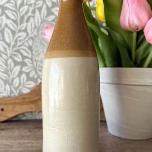 May include: A vintage stoneware bottle with a two-tone design. The top is brown, transitioning to a cream-colored body. The bottle is displayed on a wooden surface, with a bouquet of pink and yellow tulips in the background.