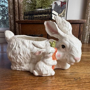 May include: A white ceramic planter shaped like a large rabbit with a smaller rabbit holding an orange carrot attached to the front. The large rabbit has pink-tinted ears and nose, while the smaller rabbit has pink-tinted ears and cheeks.