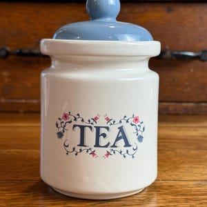 May include: A white ceramic tea canister with a removable light blue lid featuring a rounded knob. The front of the canister displays the word TEA in dark blue capital letters, surrounded by a delicate floral vine pattern in blue and pink.
