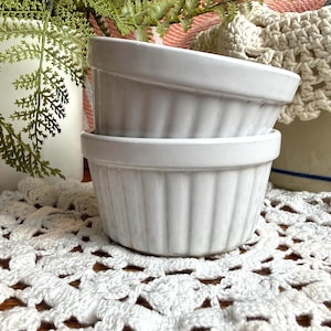 May include: Two white ceramic ramekins stacked on top of each other. Each ramekin features a classic fluted exterior design with vertical ridges and a smooth, flat rim. The glossy white finish reflects light, highlighting the cylindrical shape.